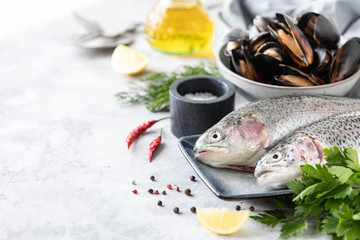 Two raw rainbow trout fish on a plate, greens and fresh vegetables for preparing healthy and tasty food. White pan with mussels on a concrete background. Healthy diet and delicious culinary concept.