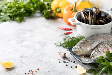 Two raw rainbow trout fish on a plate, greens and fresh vegetables for preparing healthy and tasty food. White pan with mussels on a concrete background. Healthy diet and delicious culinary concept.