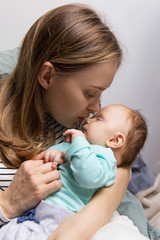 Young mother holding and kissing sleepy baby. Portrait of young woman and cute little child in home interior. Rocking baby concept