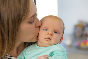 Closeup of new mom holding and kissing baby. Portrait of young woman and cute little child in home interior. Maternity or baby care concept