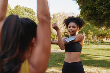 Diverse female friends standing face to face exercising together in the morning at park - fitness friends warming up their muscles together while chatting