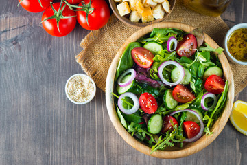 Fresh healthy salad with delicious ruccola, spinach, cabbage, arugula, feta cheese, red onion, cucumber, sesame seeds and cherry tomato on wooden background. Healthy and diet food concept.