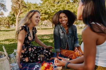 Foto op Plexiglas Group of smiling multiracial female best friends sitting together on blanket with fruits enjoying at picnic in the park - group of healthy friends having a picnic © StratfordProductions