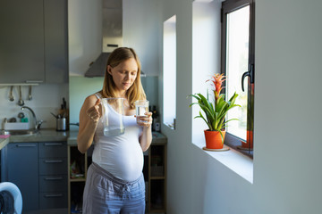 Pensive expectant mother drinking water in kitchen, holding jug and glass. Pregnant young woman spending leisure time at home. Prenatal care and thirst concept