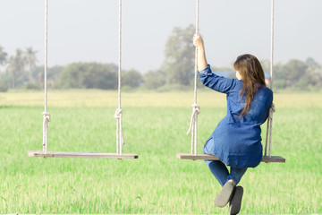 An Asian woman is happily sitting on a swing hanging among nature.  On vacation