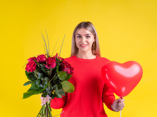 Portrait beautiful blonde young woman dressed in red holding bouquet of red roses and st. valentine heart shape balloon, look in a camera. Romantic gift concept over yellow background