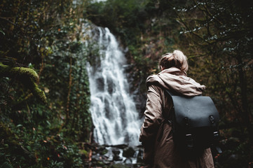 girl with a backpack on her back on a path overgrown with trees and a moss in a dark gloomy forest in the rain and looks at a high powerful waterfall - 313600158