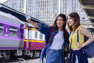 Young Asian women taking selfie photo together at train station. Happy traveler backpacker friends at railway station using smartphone take photo.