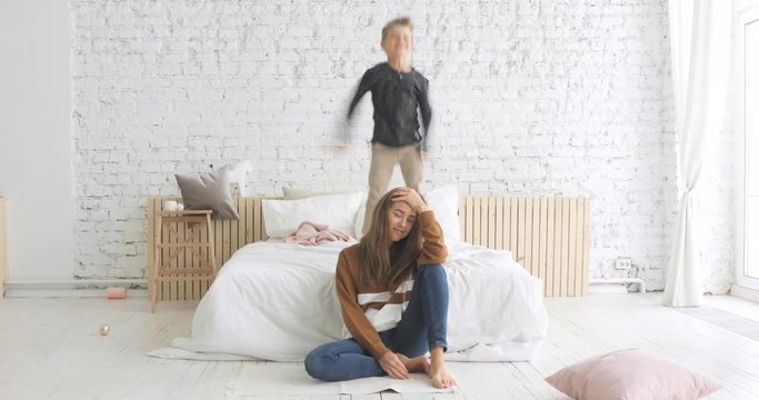 The hardships of motherhood. Hyperactive preschool son boy with attention deficit hyperactivity disorder ADHD playing jumping on bed, mom suffers lack of energy, mother tired of noisy disobedient kid.