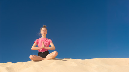 Fototapeta na wymiar ein junges Mädchen macht im Sommer Yoga auf einer Düne im gelben Sand vor blauem Himmel