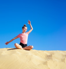 Obraz na płótnie Canvas ein junges Mädchen macht im Sommer Yoga auf einer Düne im gelben Sand vor blauem Himmel