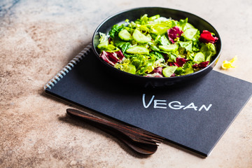 Vegan food concept. Green vegetable salad in black plate and notebook with the word vegan.