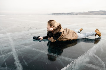 a young girl in a fur coat lies on transparent cracked ice in the middle of the lake, she is happy and smiling - 313611120