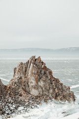 a textured lonely large rock towers over a frozen clear lake covered with blue cracked ice