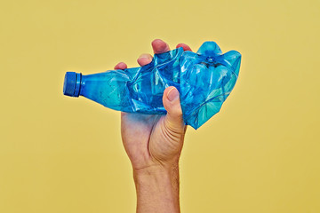 Hand squeezing blue plastic bottle on yellow background. Zero waste and consumption reduction of...