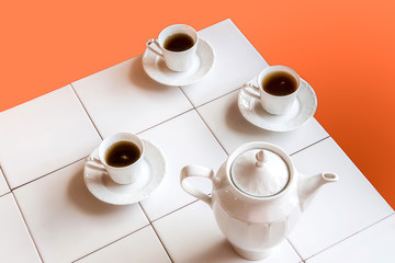 White tea cups and teapot on white tiles table over orange red background..