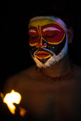 Obraz premium Face of Indian brunette man with his face illuminated and painted by vibrant colors like a tribe standing with candle light in front of a black studio copy space background. Indian hi fashion.