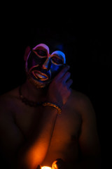 Close up of Indian brunette man with his face illuminated and painted by black and golden like a tribe standing with candle light in front of a black studio copy space background. Indian hi fashion.