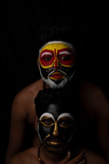 Indian brunette men with their faces painted by vibrant colors like a tribe standing in front of a black studio copy space background. Indian hi fashion.