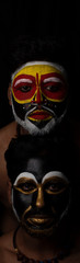 Indian brunette men with their faces painted by vibrant colors like a tribe standing in front of a black studio copy space background. Indian hi fashion.