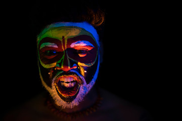 Obraz premium Face of Indian brunette man with his face illuminated and painted by vibrant colors like a tribe standing with candle light in front of a black studio copy space background. Indian hi fashion.