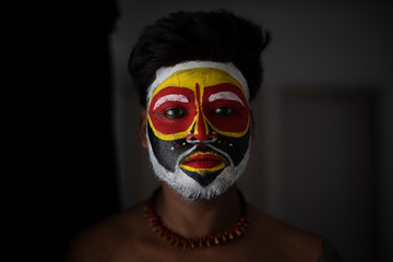 Close up portrait of an Indian brunette man with his face painted by vibrant colors like a tribe standing in front of a studio background. Indian hi fashion.