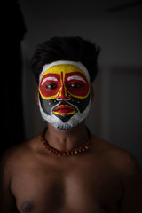 Face of Indian brunette man with his face illuminated and painted by vibrant colors like a tribe standing with candle light in front of a black studio copy space background. Indian hi fashion.