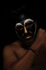 Close up of Indian brunette man with his face illuminated and painted by black and golden like a tribe standing with candle light in front of a black studio copy space background. Indian hi fashion.