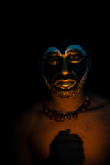 Fototapeta na wymiar Close up of Indian brunette man with his face illuminated and painted by black and golden like a tribe standing with candle light in front of a black studio copy space background. Indian hi fashion.