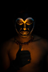Close up of Indian brunette man with his face illuminated and painted by black and golden like a tribe standing with candle light in front of a black studio copy space background. Indian hi fashion.