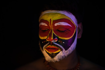 Face of Indian brunette man with his face illuminated and painted by vibrant colors like a tribe standing with candle light in front of a black studio copy space background. Indian hi fashion.