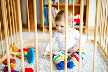 Beautiful little baby girl standing inside playpen. Cute adorable child playing with colorful toys....