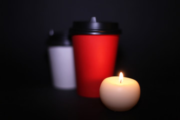 Red and white paper Cup for coffee and cappuccino stands on a black background and next to them a candle, evening and romantic atmosphere.