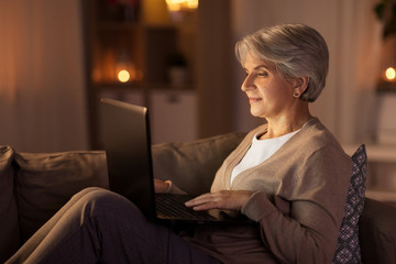 technology, old age and people concept - happy senior woman with laptop computer at home in evening