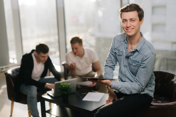 Focused young man uses a tablet and looking at the camera in sunlit office. Two young startup businessman work together behind a laptop in the background.