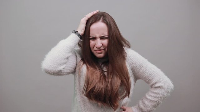 Unhealthy caucasian lady keeping hand on head, having headache, keeping hand on belt wearing blue sweater isolated on gray background in studio. People emotions, lifestyle concept.