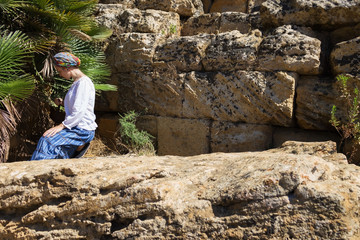 Woman in a white blouse, a long skirt and a turban. Woman sits on antique ruins near palm tree. The Valle dei Templi, Agrigento, Sicily, Italy