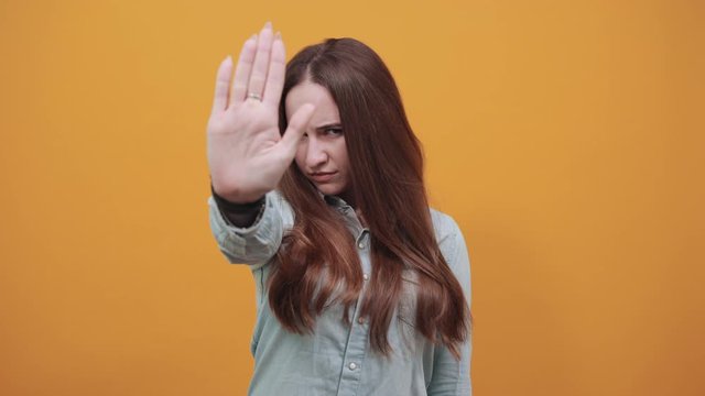 Serious caucasian brunette woman in denim blue shirt showing palm at camera, doing stop gesture isolated on orange background in studio. People emotions, lifestyle concept.
