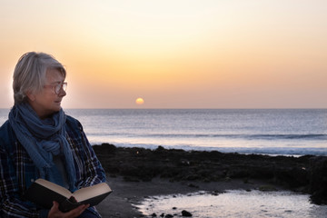 A senior woman relax sitting on the beach with a book. Sunset at the horizon over water. Seascape. One people with gray hair and eyeglasses