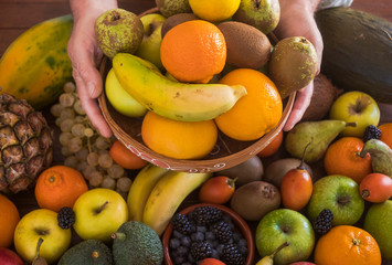 Two hands of a person hold a basket full of fruit. Background with an assortment of freshly picked fresh fruit. Many vitamins together on a wooden table.