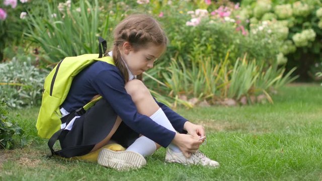 A schoolgirl with a backpack sits on a lawn and learns to tie shoelaces. Preparation for school.