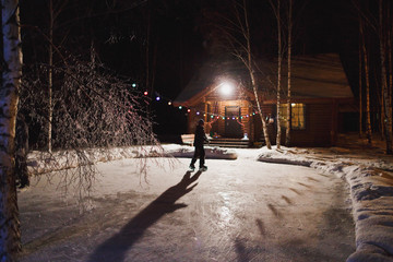 Young boy in winter clothes skating on a skating rink decorated with lanterns at the house in the countryside on a winter evening. Selective focus. Winter leisure activity.