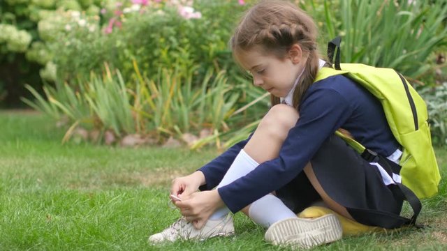 A schoolgirl with a backpack sits on a lawn and learns to tie shoelaces. Preparation for school.