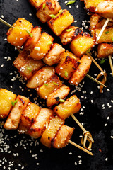 Grilled skewers with pineapple fruit and chicken meat  sprinkled with sesame seeds and spices on a black background, close-up . Fruit and meat skewers
