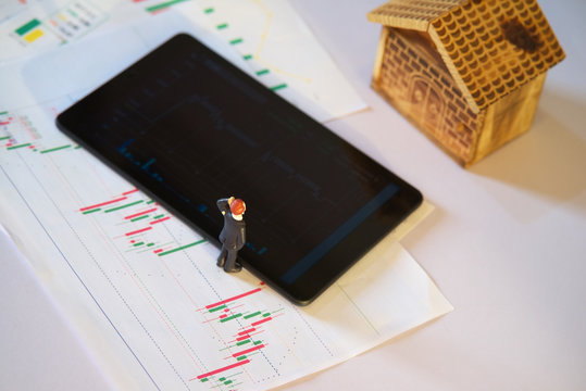 miniature business man with stock exchange graph data on white paper and blurred tablet with blurred wooden house, business or stock exchange graph analysis concept
