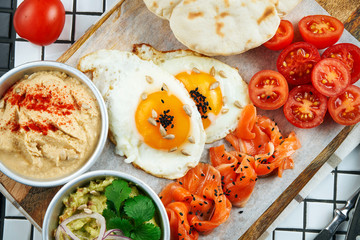 Unusual breakfast. Fried eggs, cherry tomatoes, guacamole hummus and salmon with pita on a board on a white background. Top view food.