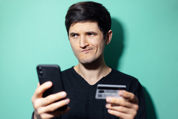 Studio portrait of young surprised man with smartphone and credit card in hand on background of aqua menthe color.