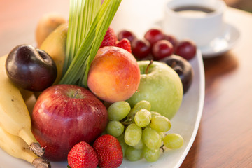 Plate of colorful various fruits with white cup of coffee in the back over a wood table with natural light