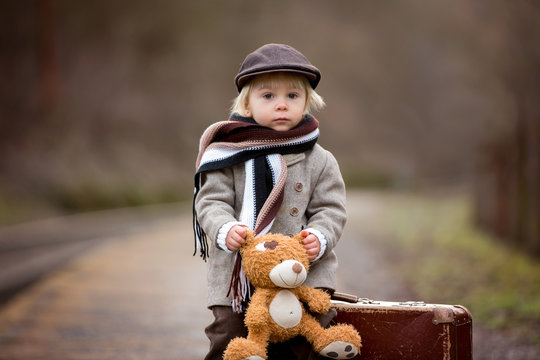 Adorable boy on a railway station, waiting for the train with suitcase and sweet teddy bear