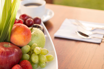Colorful various fruits with white cup of coffee and spoon and fork in the back over a wood table with natural light